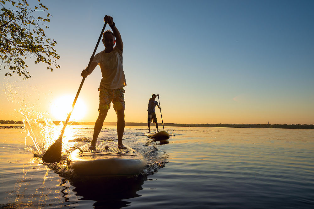 Men, friends sail on a SUP boards in a rays of rising sun. Stand up paddle boarding - awesome active recreation in nature. Backlight.
