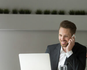 Smiling businessman wearing suit sitting at working desk using typing on laptop, making answering call, talking on the phone, consulting client about email by cell, holding mobile interview in office