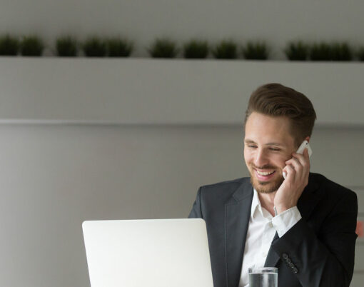 Smiling businessman wearing suit sitting at working desk using typing on laptop, making answering call, talking on the phone, consulting client about email by cell, holding mobile interview in office