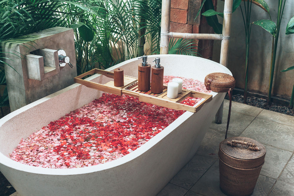 Bathtub with flower petals and beauty products on wooden tray. Organic spa relaxation in luxury Bali outdoor bath.