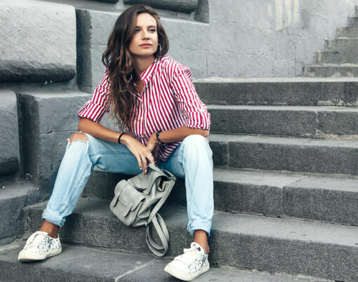 Fashion model wearing ripped boyfriend jeans, red striped shirt, sneakers and backpack posing in the city street. Fashion urban outfit. Casual everyday clothing style.