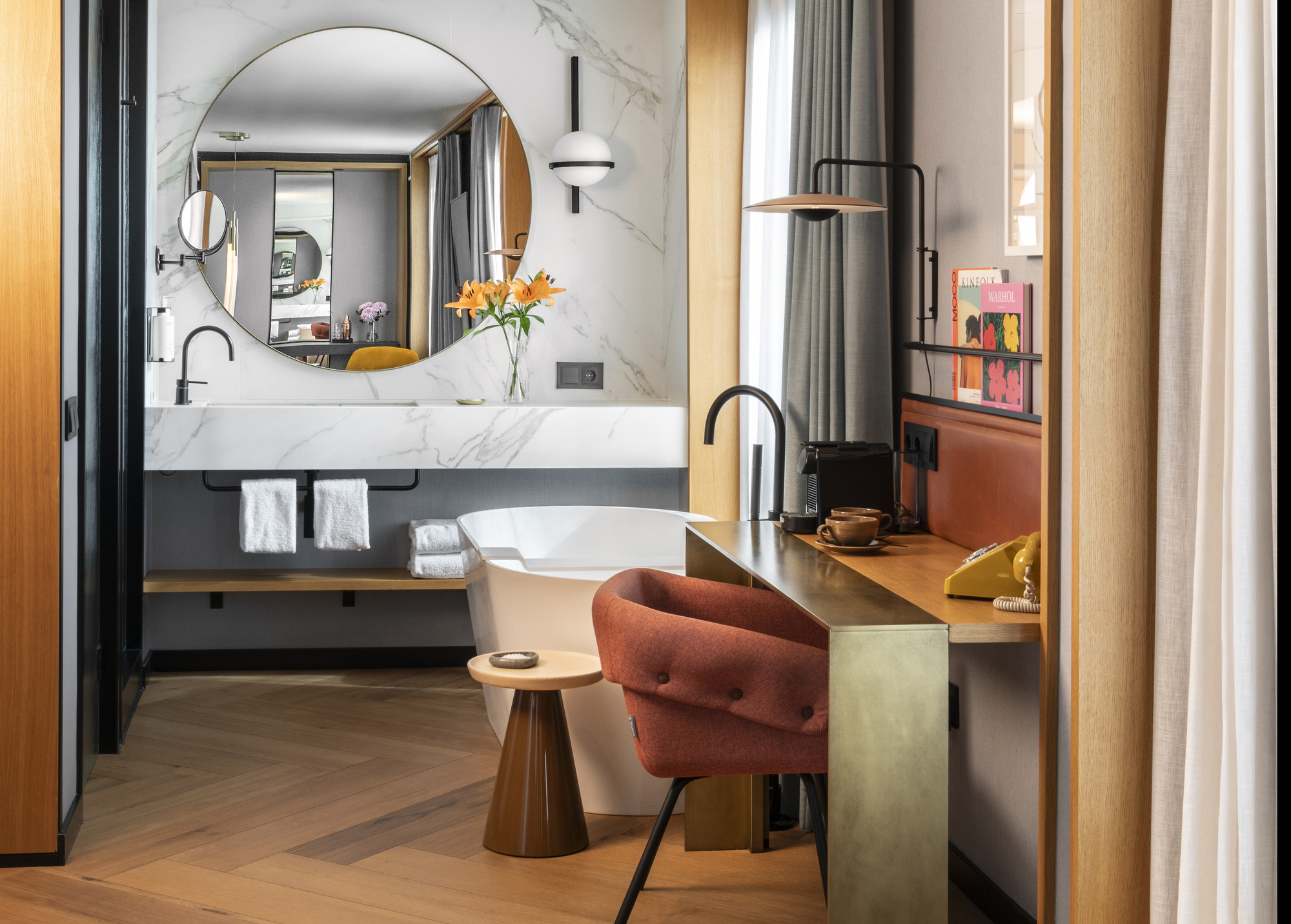 Luxurious bathroom with a round marble vanity mirror, vase of orange lilies, and a freestanding tub beside a marble wall.